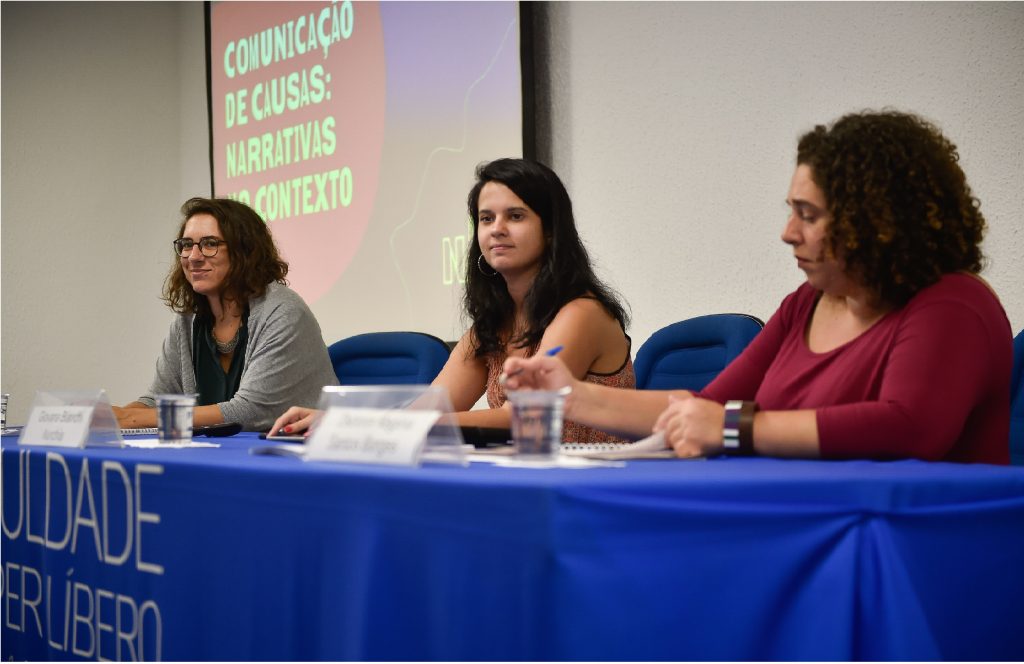 Giovana Bianchi, Laura Leal, Débora Borges estão sentadas em uma bancada. Ao fundo está projetado em um painel: comunicação de causas: narrativas 